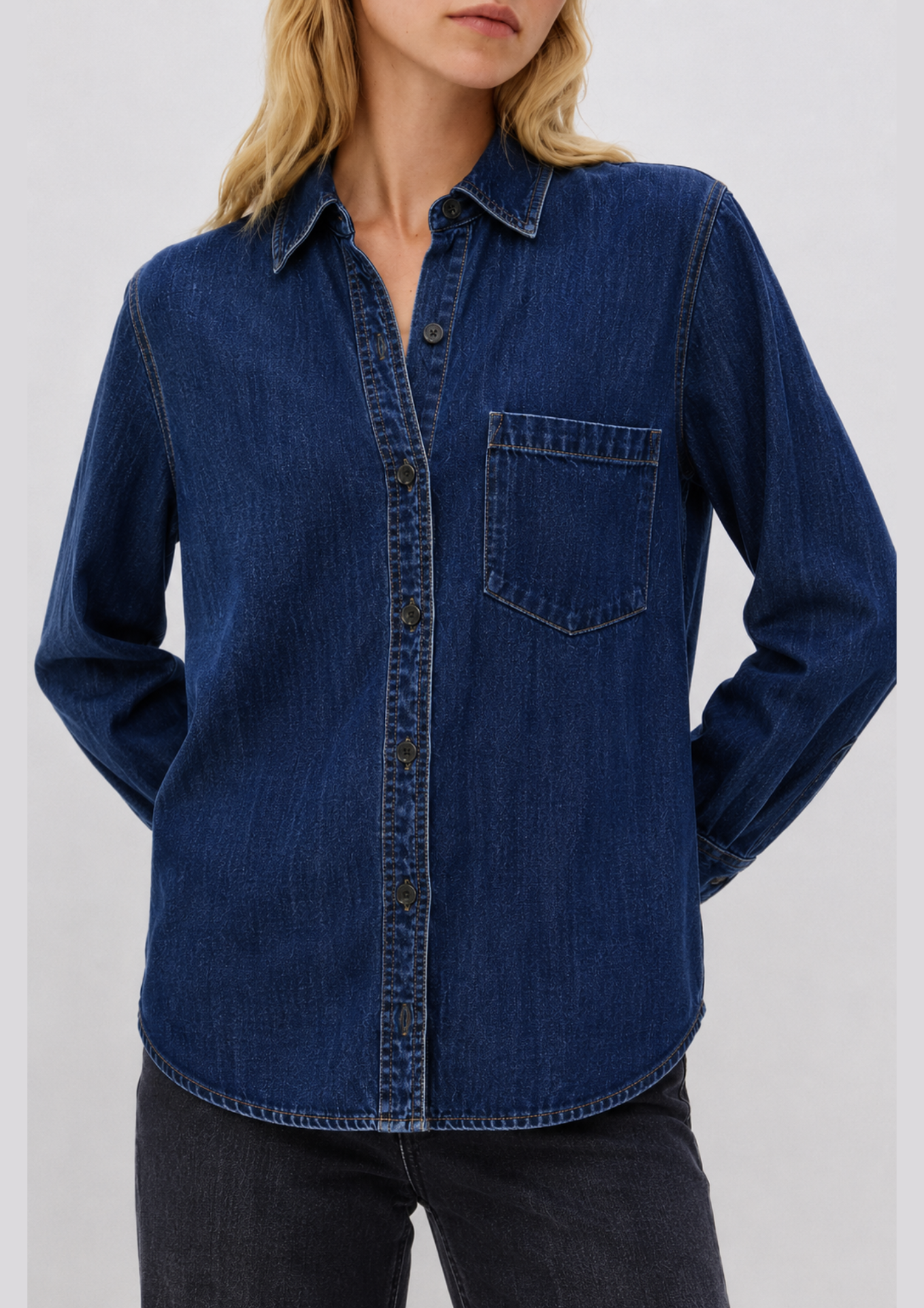 A woman wearing a dark blue denim long-sleeve button-up shirt with a chest pocket, paired with dark jeans, against a plain light gray background.