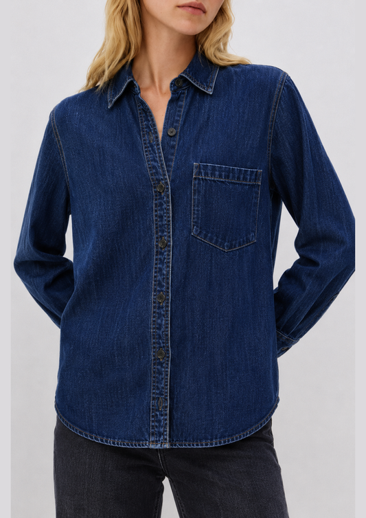 A woman wearing a dark blue denim long-sleeve button-up shirt with a chest pocket, paired with dark jeans, against a plain light gray background.