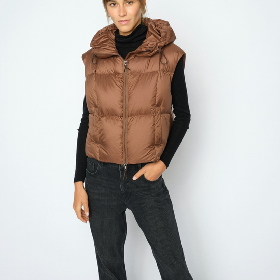 A woman modeling a brown hooded puffer vest over a black long-sleeve top and black jeans, standing against a light gray studio background.