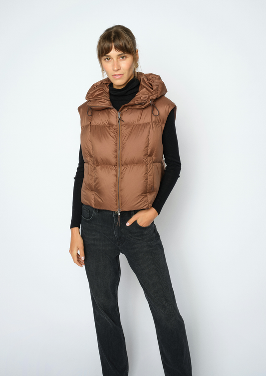 A woman modeling a brown hooded puffer vest over a black long-sleeve top and black jeans, standing against a light gray studio background.