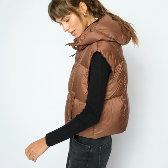A woman wearing a brown puffer vest over a black long-sleeve shirt and dark gray jeans, photographed against a plain light gray studio background.