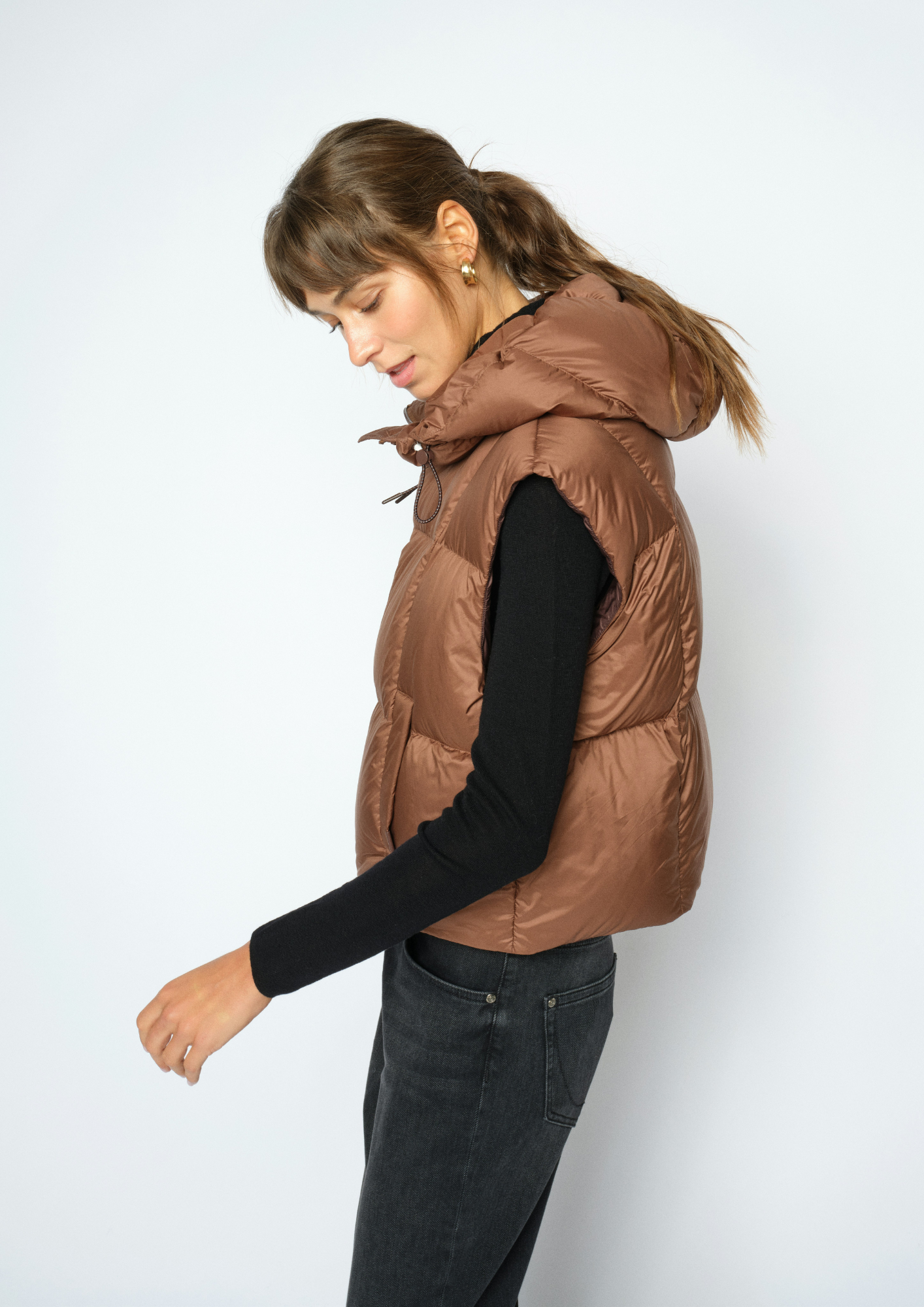 A woman wearing a brown puffer vest over a black long-sleeve shirt and dark gray jeans, photographed against a plain light gray studio background.