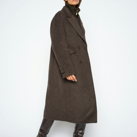 A model wearing a long, dark brown double-breasted wool coat over a black turtleneck, paired with black leather boots, photographed against a light gray studio background.