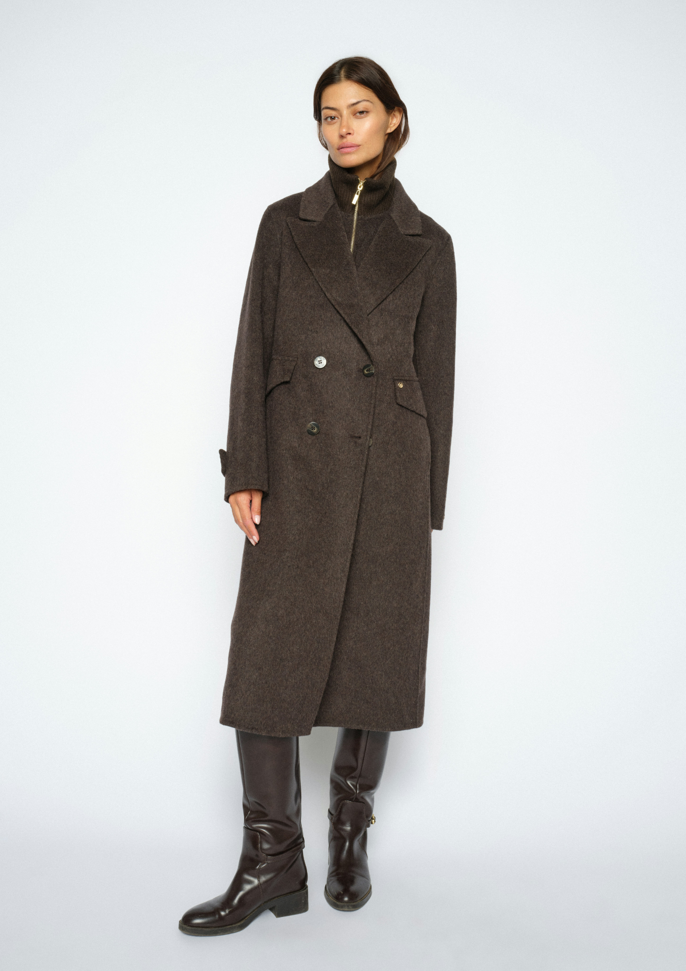 A model wearing a dark brown double-breasted wool coat with a high collar, paired with dark brown leather boots, against a light gray studio background.