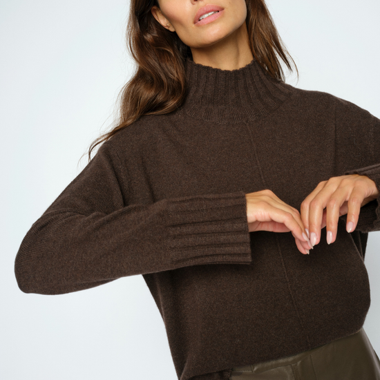 A woman wearing a dark brown ribbed mock neck sweater and dark brown leather pants, posing against a light gray studio background.
