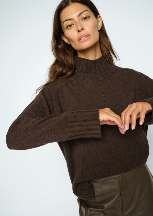A woman wearing a dark brown ribbed mock neck sweater and dark brown leather pants, posing against a light gray studio background.