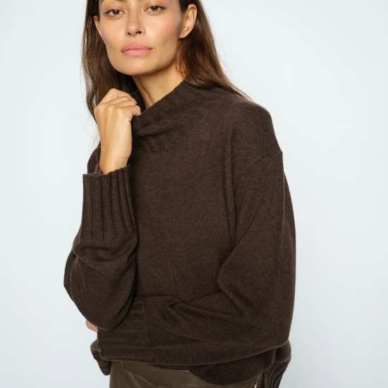 A woman modeling a dark brown knit sweater with a mock neck and ribbed cuffs, paired with dark brown leather pants, against a light gray studio background.