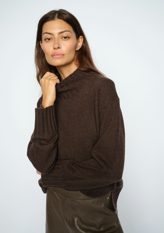 A woman modeling a dark brown knit sweater with a mock neck and ribbed cuffs, paired with dark brown leather pants, against a light gray studio background.