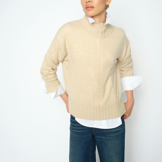 A woman modeling a beige mock-neck knit sweater layered over a white collared shirt and dark blue jeans, photographed against a clean white studio background.