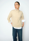A woman modeling a beige mock-neck knit sweater layered over a white collared shirt and dark blue jeans, photographed against a clean white studio background.