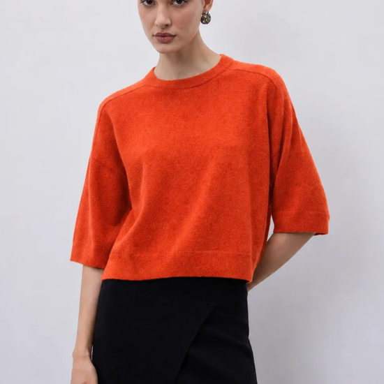 A woman modeling a bright orange short-sleeve knit sweater paired with a black skirt, photographed against a plain light gray studio background.