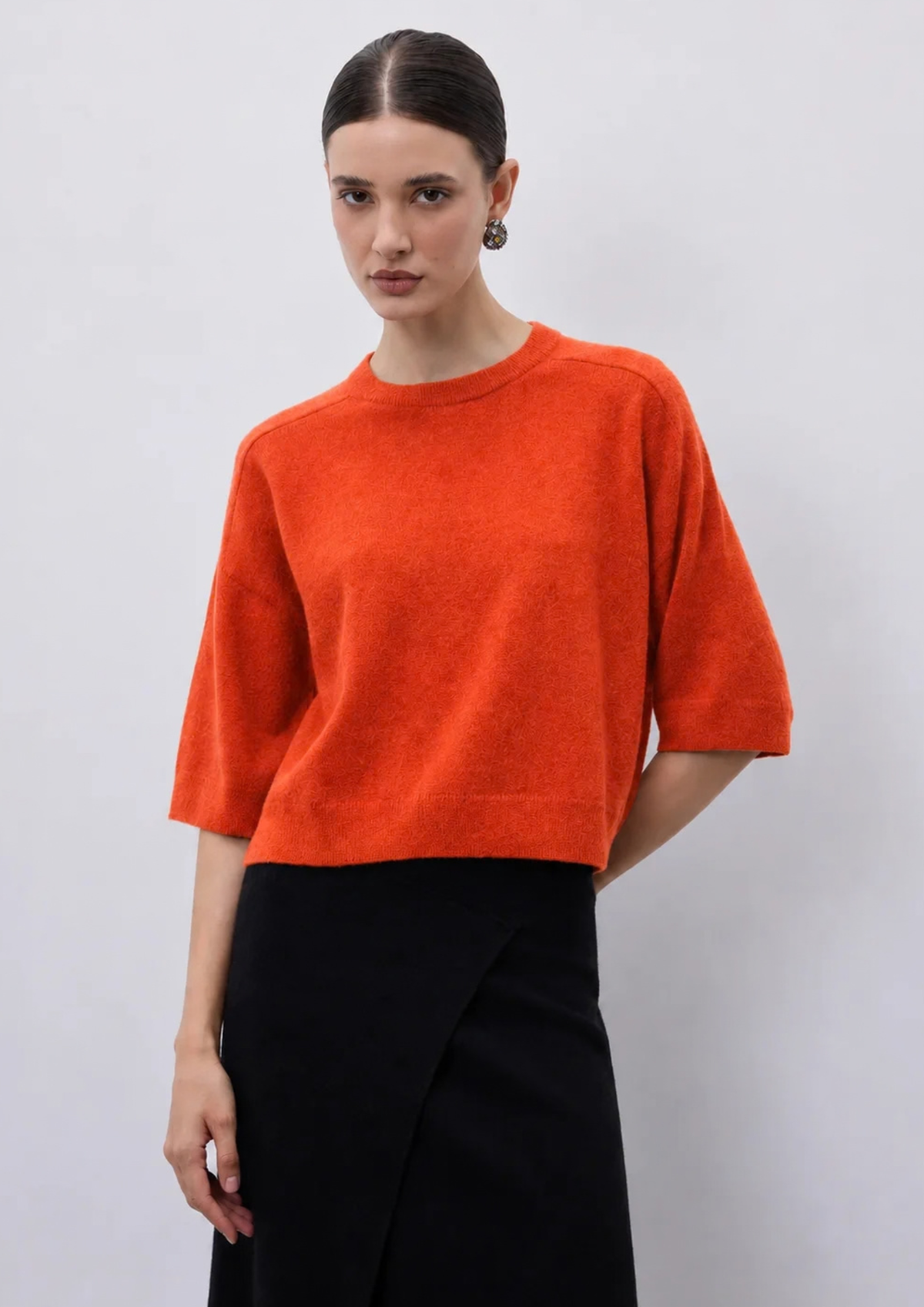 A woman modeling a bright orange short-sleeve knit sweater paired with a black skirt, photographed against a plain light gray studio background.