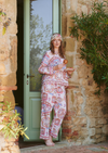 A woman stands barefoot in front of a rustic stone wall and green door, wearing a matching pajama set with a red, blue, and cream paisley pattern and a matching headscarf. She holds a small plate with a drink.