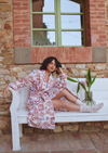 A woman sits on a white bench outdoors against a brick and stone wall, wearing a long-sleeve patterned robe in pink, red, and white. A glass vase with flowers sits beside the bench.