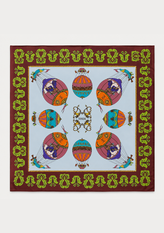 A square silk scarf featuring a colorful hot air balloon pattern on a light blue background, surrounded by a brown border with green and gold decorative motifs.