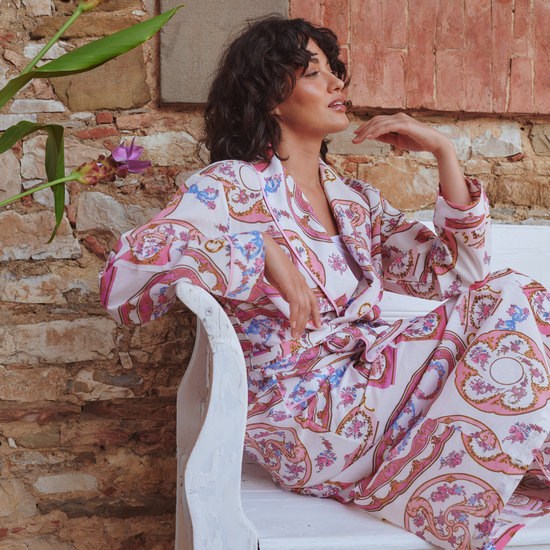 A woman wearing a long-sleeve pajama set with a pink, red, and blue paisley pattern sits on a white wooden bench against a rustic brick wall.