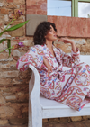 A woman wearing a long-sleeve pajama set with a pink, red, and blue paisley pattern sits on a white wooden bench against a rustic brick wall.