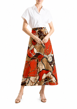 A model wearing a white short-sleeve button-up shirt and a high-waisted midi skirt with an abstract chain-link print in red, beige, and brown, paired with beige high-heeled sandals against a white background.