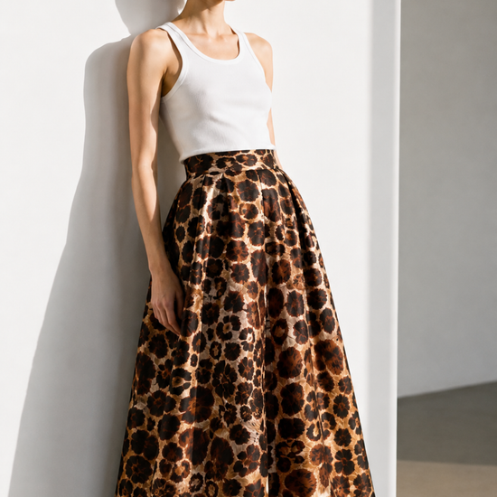 A model wearing a white tank top and a high-waisted, full-length leopard-print skirt with a glossy satin-like finish, leaning against a white wall in a minimalist studio setting.