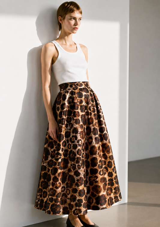 A model wearing a white tank top and a high-waisted, full-length leopard-print skirt with a glossy satin-like finish, leaning against a white wall in a minimalist studio setting.