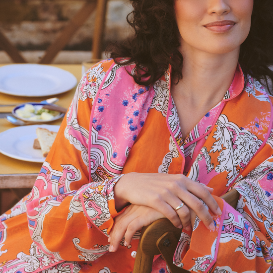 A woman with curly dark hair wearing a vibrant orange and pink paisley patterned robe, sitting at a wooden table with plates in a warm, rustic setting.