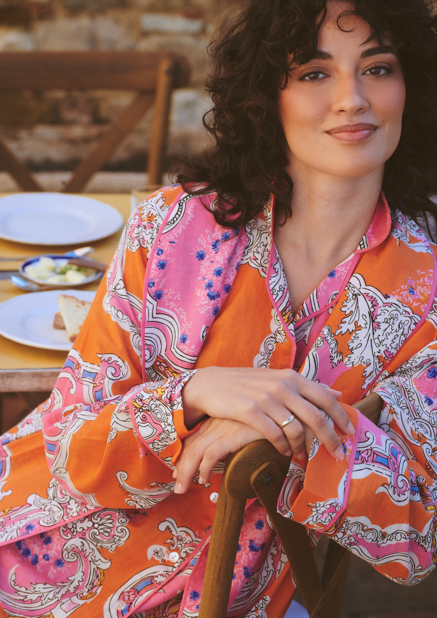 A woman with curly dark hair wearing a vibrant orange and pink paisley patterned robe, sitting at a wooden table with plates in a warm, rustic setting.