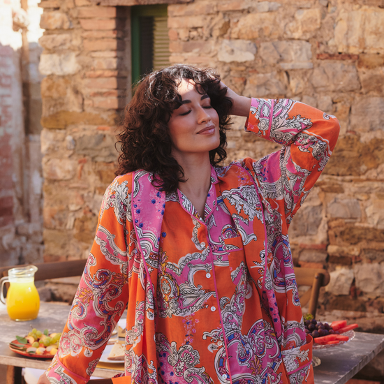 A woman wearing a vibrant orange and pink paisley patterned long-sleeve shirt stands outdoors in front of a rustic stone wall, with a wooden table and a pitcher of orange juice visible in the background.