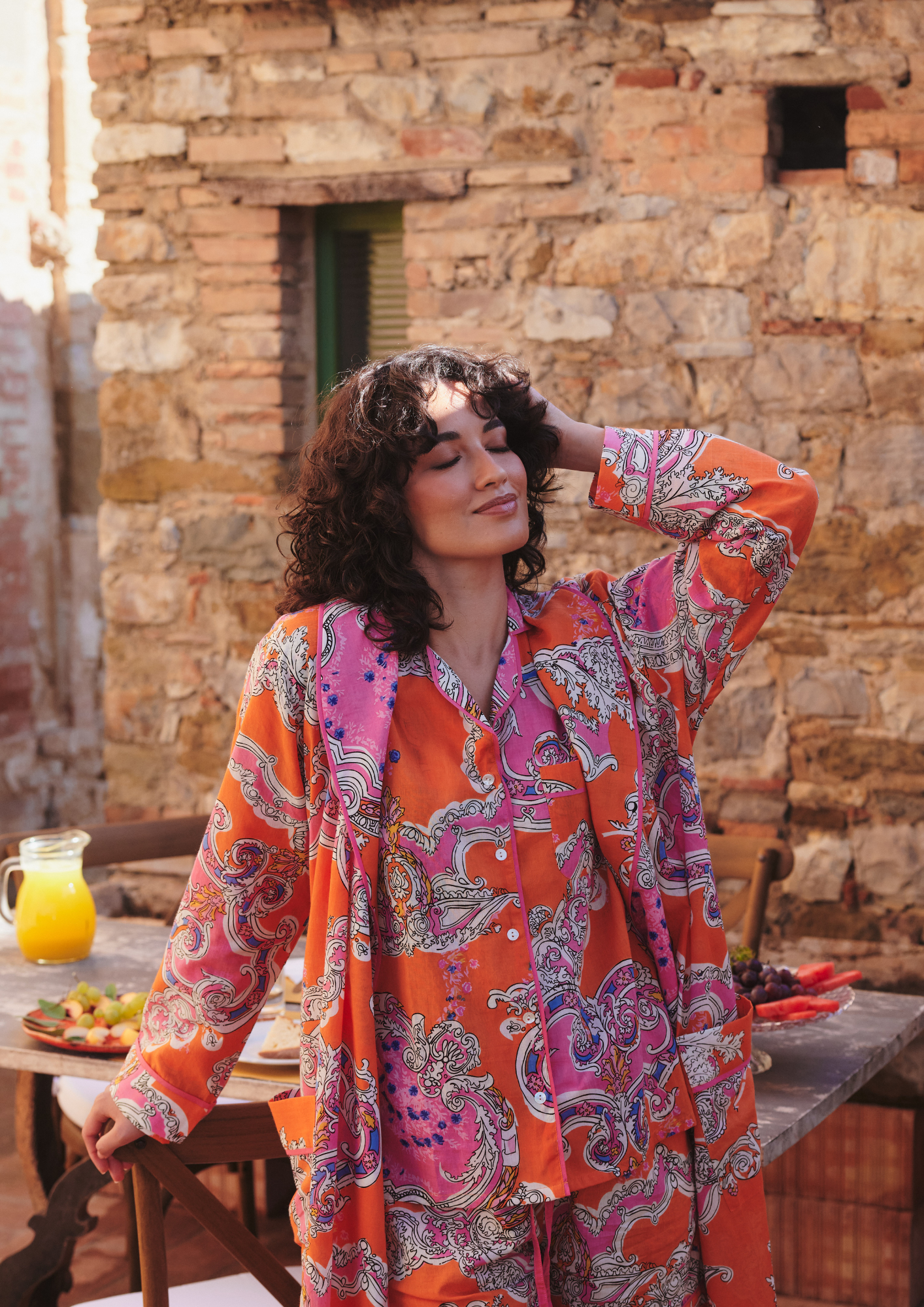 A woman wearing a vibrant orange and pink paisley patterned long-sleeve shirt stands outdoors in front of a rustic stone wall, with a wooden table and a pitcher of orange juice visible in the background.