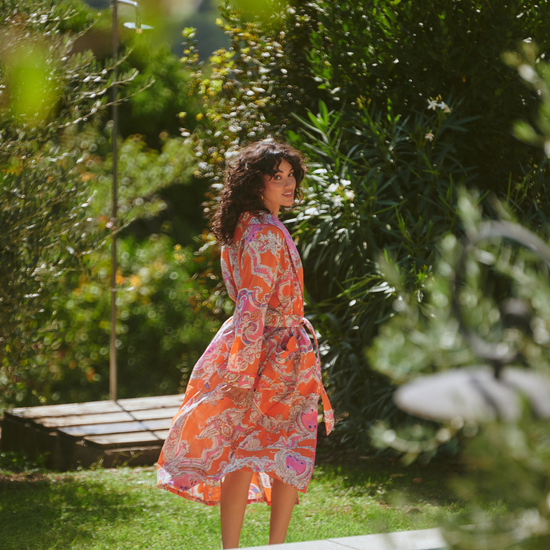 A woman wearing a long-sleeved orange and pink paisley maxi dress stands by a swimming pool in a lush, green outdoor garden setting.