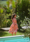 A woman wearing a long-sleeved orange and pink paisley maxi dress stands by a swimming pool in a lush, green outdoor garden setting.