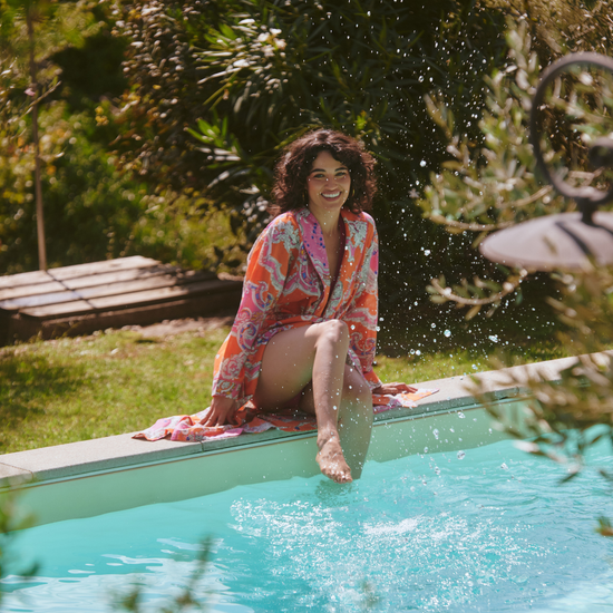 A woman sitting on the edge of a swimming pool in a lush garden, wearing a colorful patterned robe with her legs dangling into the turquoise water.