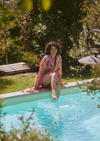 A woman sitting on the edge of a swimming pool in a lush garden, wearing a colorful patterned robe with her legs dangling into the turquoise water.