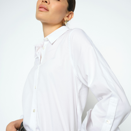 A woman wearing a white long-sleeve button-down shirt with three-quarter puffed sleeves, paired with a black leather belt and dark jeans, photographed against a clean white studio background.