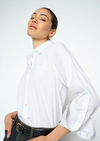 A woman wearing a white long-sleeve button-down shirt with three-quarter puffed sleeves, paired with a black leather belt and dark jeans, photographed against a clean white studio background.