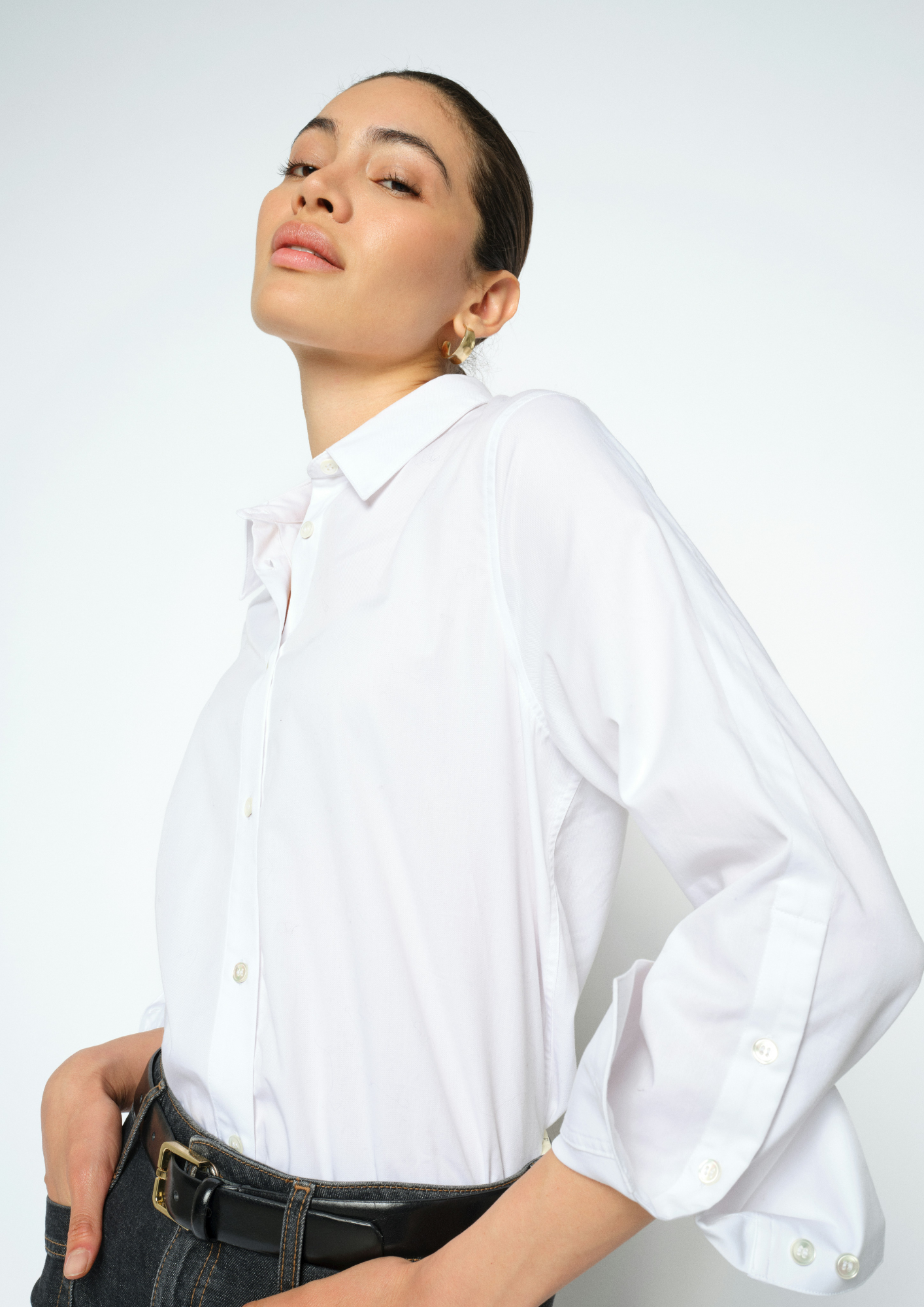 A woman wearing a white long-sleeve button-down shirt with three-quarter puffed sleeves, paired with a black leather belt and dark jeans, photographed against a clean white studio background.