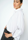 A woman wearing a white long-sleeve button-up shirt tucked into dark jeans, shown from a side-back angle against a plain white studio background.