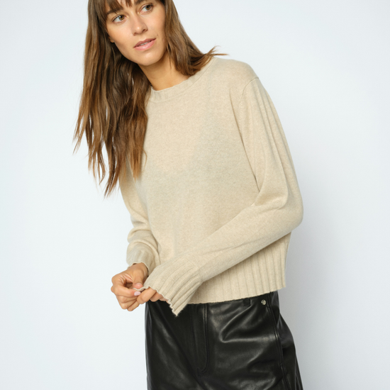 A woman wearing a light beige knit sweater and black leather pants, posing against a light gray studio background.