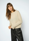 A woman wearing a light beige knit sweater and black leather pants, posing against a light gray studio background.
