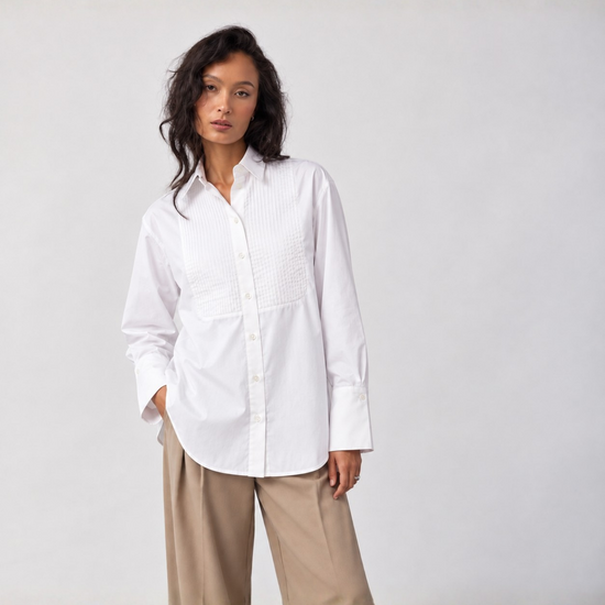 A woman modeling a white long-sleeve button-up shirt with textured vertical pleats on the chest, paired with beige wide-leg trousers, against a plain light gray studio background.