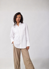 A woman modeling a white long-sleeve button-up shirt with textured vertical pleats on the chest, paired with beige wide-leg trousers, against a plain light gray studio background.