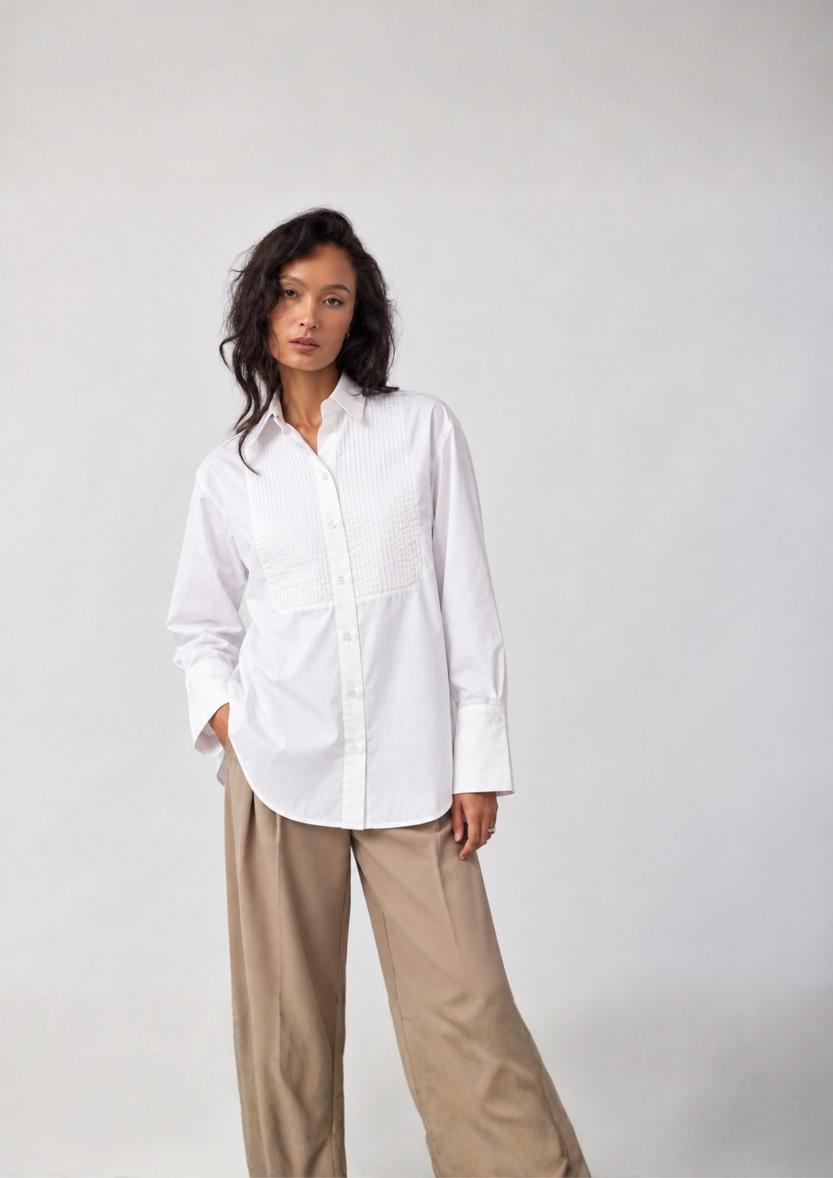 A woman modeling a white long-sleeve button-up shirt with textured vertical pleats on the chest, paired with beige wide-leg trousers, against a plain light gray studio background.