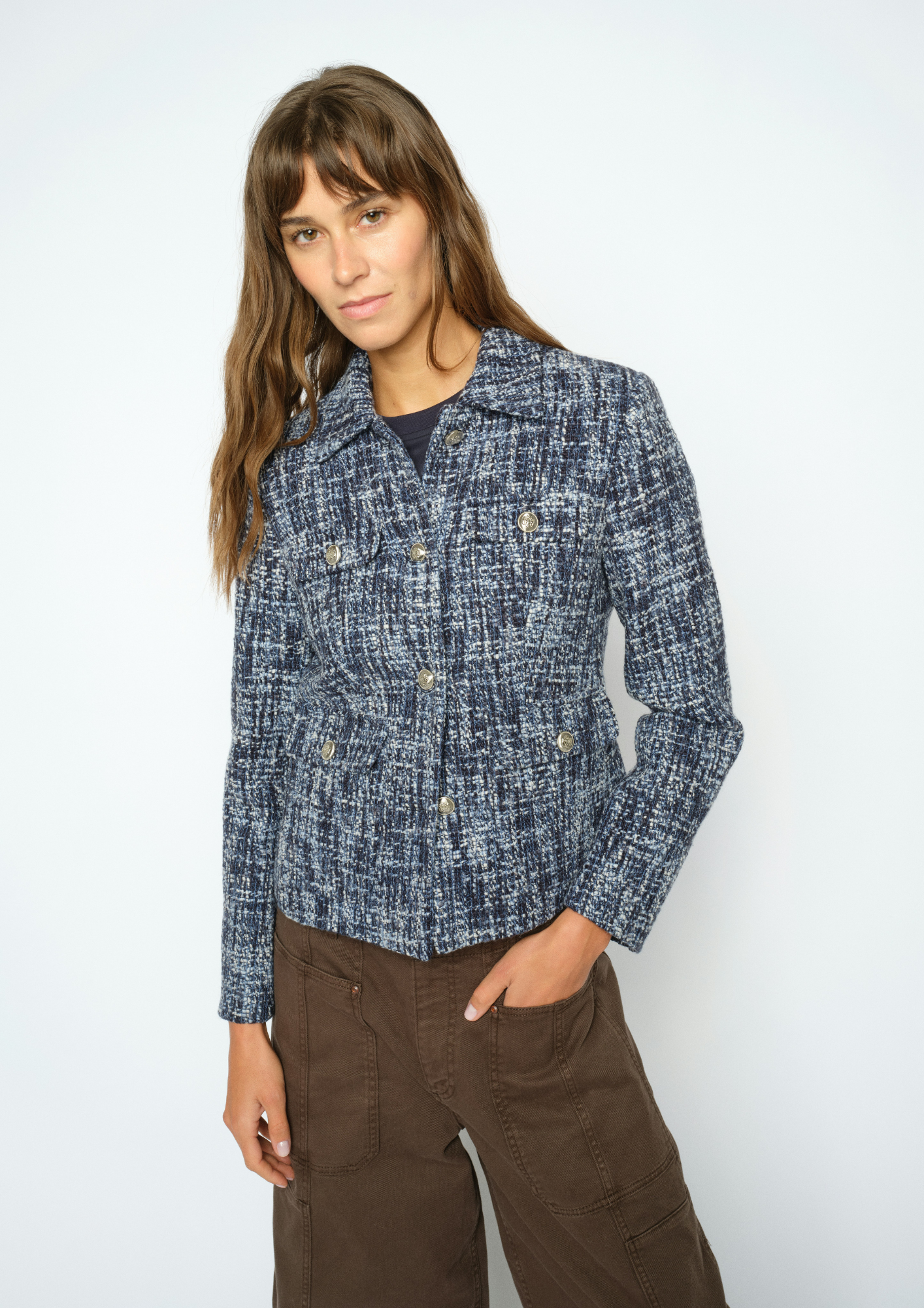 A woman modeling a blue and white textured tweed jacket with brown pants against a plain white studio background.