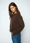 A woman wearing a brown knit crewneck sweater and blue jeans, standing against a light gray studio background.