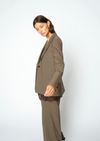 A woman modeling a matching taupe brown two-piece suit, featuring a structured blazer and wide-leg trousers, posed against a light gray studio background.