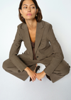 A woman wearing a tailored taupe-brown blazer and matching wide-leg trousers, seated cross-legged against a plain light gray studio background.