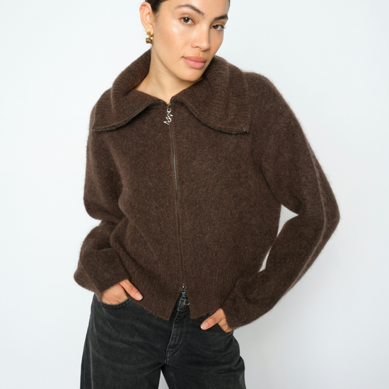 A woman wearing a brown fuzzy zip-up jacket with a wide collar and dark wide-leg jeans, standing against a plain light gray background.