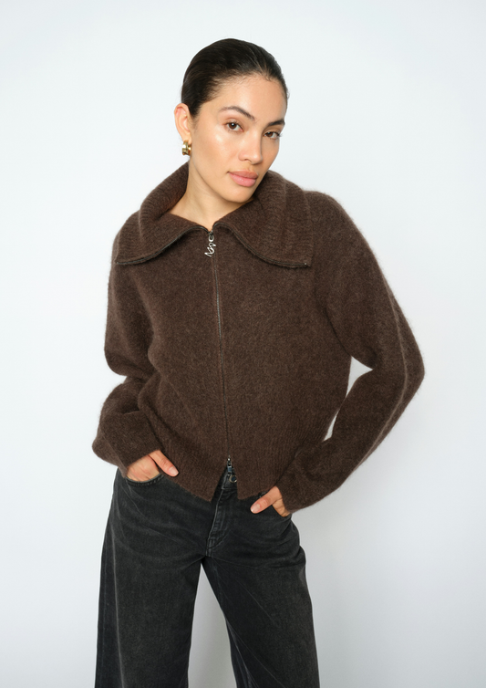 A woman wearing a brown fuzzy zip-up jacket with a wide collar and dark wide-leg jeans, standing against a plain light gray background.