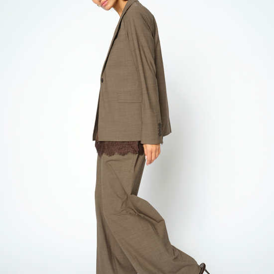 A woman modeling a matching taupe brown wide-leg trouser suit with an oversized blazer, featuring a dark lace underlayer at the hem and dark brown pointed-toe heels, set against a plain light gray studio background.