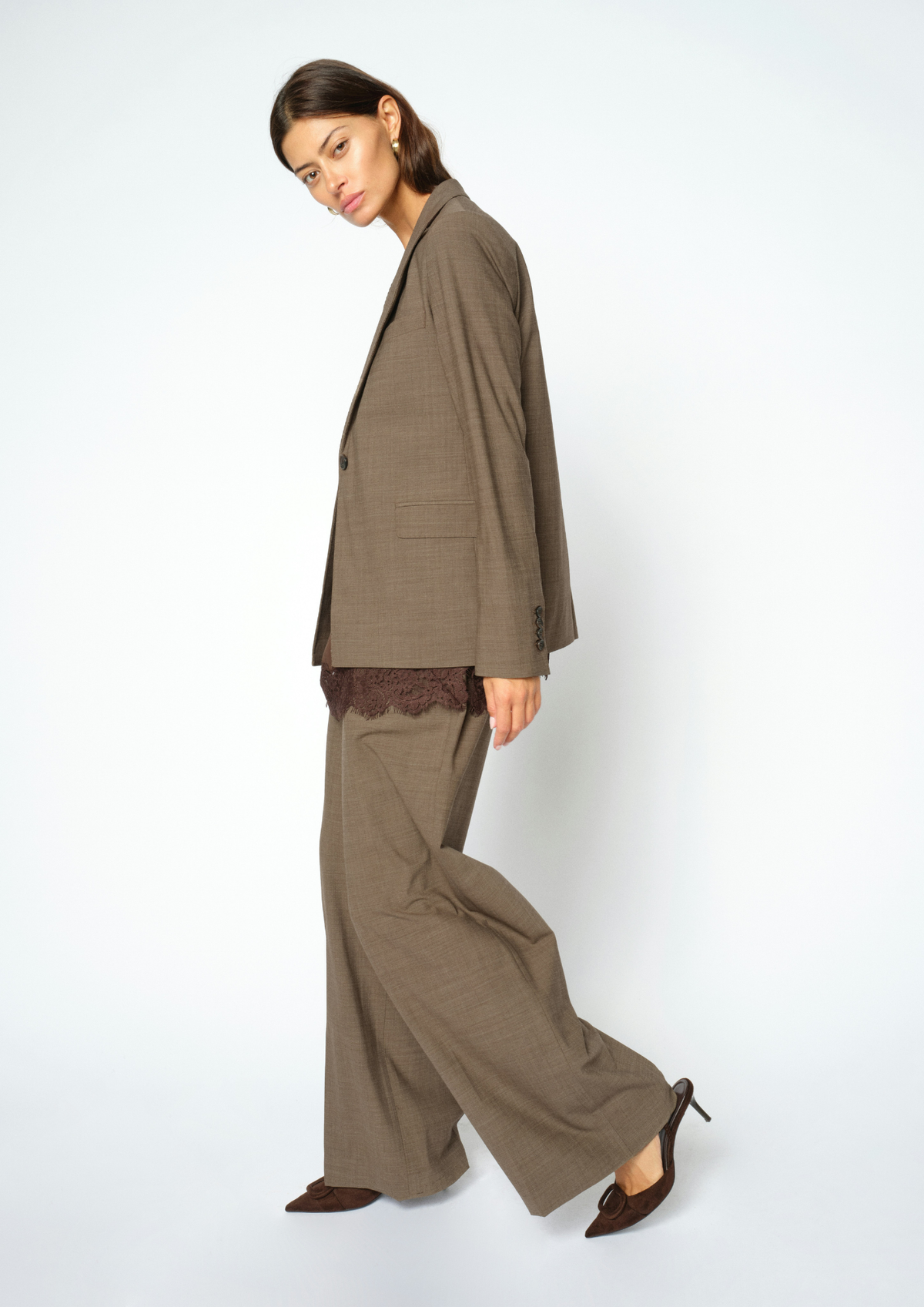 A woman modeling a matching taupe brown wide-leg trouser suit with an oversized blazer, featuring a dark lace underlayer at the hem and dark brown pointed-toe heels, set against a plain light gray studio background.