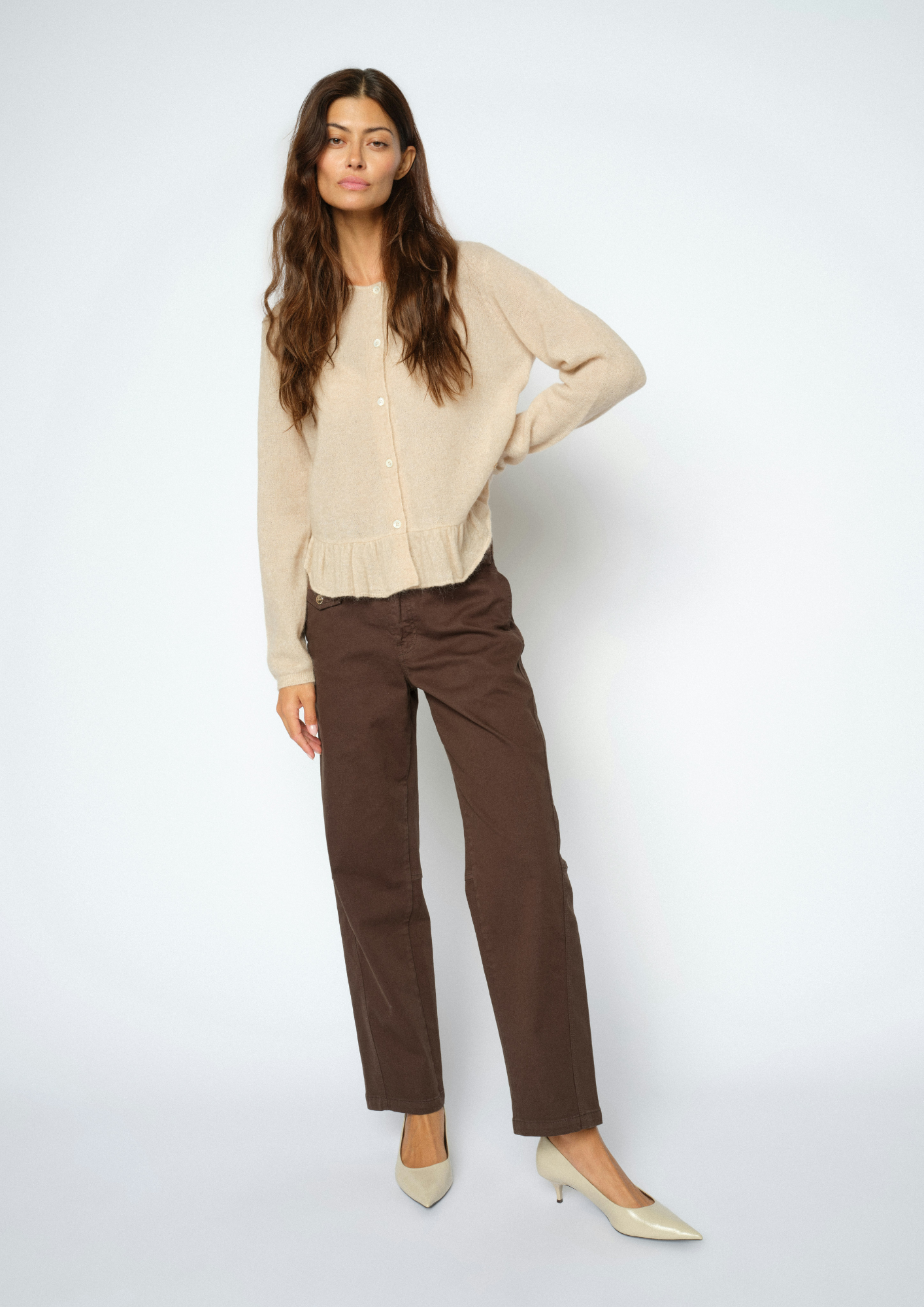 A woman modeling a beige knit cardigan sweater and brown straight-leg trousers, paired with beige pointed-toe heels, against a light gray studio background.